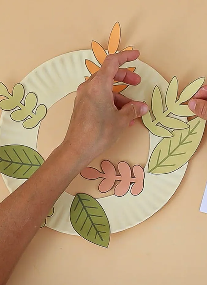 Paper plate leaf wreath being decorated with paper leaves by a child
